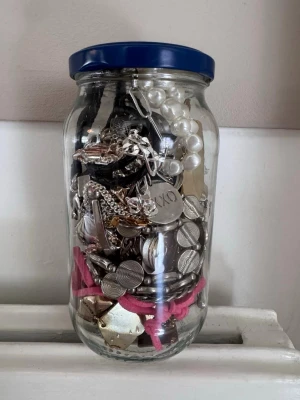 Stor burk med blandade smycken - En glasburk fylld med olika smycken som armband, halsband och ringar i blandade färger och material. Innehåller både metall, plast och textil. Perfekt för dig som vill ha unika accessoarer till din stil.