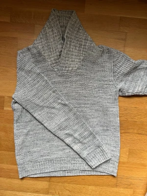 Grå stickad polotröja från H&M - Säljer en grå stickad polotröja från H&M med lång ärm och bred, ribbad krage. Tröjan har en klassisk, ribbad struktur och är perfekt för kyligare dagar. Enkel att matcha till olika outfits och har en neutral, ljusgrå ton.