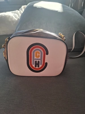 Coach crossbody handväska med logga - Snygg crossbody handväska från Coach i vitt och beige med färgglad CH-logga på framsidan. Väskan har guldfärgade detaljer, dragkedja och justerbar axelrem. Insidan är fodrad i vinrött tyg och har en innerficka. Perfekt för dig som gillar trendiga accessoarer. Ej äkta men good condition