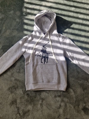 Grå hoodie med svart tryck - Snygg grå hoodie med stor svart tryck på bröstet som föreställer en polospelare och text. Hoodien har huva med vita snören och en klassisk känguruficka framtill. Perfekt för en avslappnad och trendig stil.