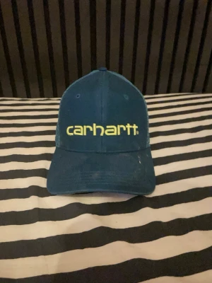 Blå Carhartt keps med mesh - Snygg blå keps från Carhartt med gul logga framtill. Kepsen har klassisk böjd skärm och mesh-paneler på sidorna för extra ventilation. Justerbar baktill för perfekt passform. Tillverkad i bomull och polyester.