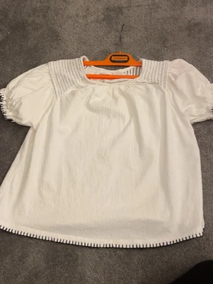 Vit blus med puffärm och broderidetaljer - Säljer en vit blus med puffiga korta ärmar och dekorativa broderade detaljer i svart vid ärmslut och nederkant. Blusen har en fyrkantig ringning och struktur i tyget, vilket ger en trendig och luftig känsla. Perfekt till jeans eller kjol för en fräsch look.
