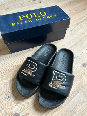 Svarta sandaler Polo Ralph Lauren - Svarta slides från Polo Ralph Lauren med stort P och tigerbrodyr på remmen. Tillverkade i syntetmaterial med bekväm innersula och klassisk design. Perfekta för chill dagar och enkel att matcha med streetwear.