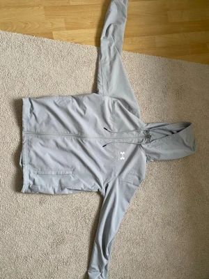 Rival Windbreaker, Under Armour, Grå - Säljer en snygg Under Armour jacka i grå färg. Stilren design med huva, dragkedja framtill och praktiska sidofickor. Perfekt för träning, vardag eller hösten. Jackan är i fint skick och redo för sin nästa ägare. Storlek M