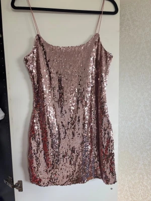Rosa paljettklänning med smala band - Glittrig rosa kortklänning täckt av paljetter och med tunna axelband. Klänningen har en rak passform och är perfekt för dig som vill sticka ut. Materialet är troligtvis polyester och den har en snygg, festlig look.
