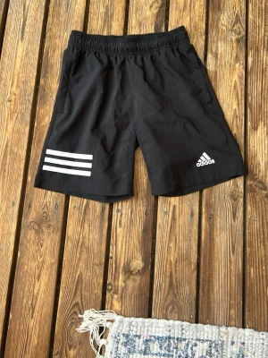 Svarta Adidas shorts med vita ränder - Säljer ett par svarta Adidas shorts med tre vita ränder på ena benet och Adidas-logga på andra. De har elastisk midja och är tillverkade i ett lätt och bekvämt material, perfekta för träning eller chill. Klassisk sportig look.