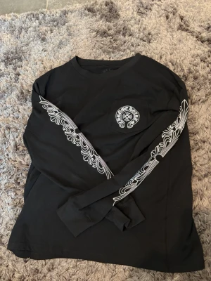 Svart långärmad chrome hearts topp med tryck - Svart långärmad chrome hearts toppmed vit dekorativt tryck på bröstfickan och längs ärmarna. Rund halsringning och avslappnad passform. Materialet ser ut att vara mjukt bomullstyg. Perfekt för en streetwear-look.
