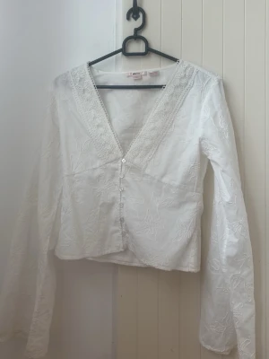 Vit blus med spetsdetaljer - Söt vit blus med broderade blommor och spets längs den djupa V-ringningen. Blusen har knäppning framtill och långa ärmar. Perfekt för en romantisk och bohemisk stil. Materialet känns lätt och luftigt.