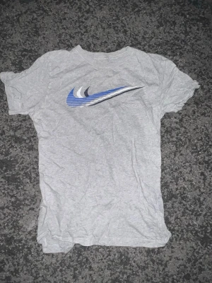 Grå t-shirt med blå Nike-logga - Säljer en grå t-shirt med en stor blå och vit Nike-logga på bröstet. T-shirten har klassisk passform och är kortärmad, perfekt till vardags. Tillverkad i mjuk bomull för skön känsla. Enkel och stilren design som funkar till det mesta.