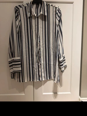 Randig skjorta från Gina Tricot - Säljer en snygg skjorta från Gina Tricot med vertikala ränder i blått, beige och vitt. Skjortan har klassisk krage, långa ärmar och knappar framtill. Perfekt för dig som gillar stilrena och trendiga plagg.