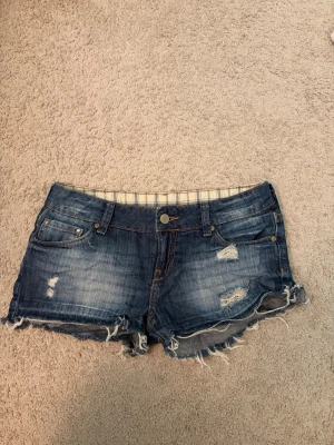 Blå slitna jeansshorts med fransar - Säljer ett par blå jeansshorts med slitna detaljer och fransig kant. Shortsen har fem fickor, knapp och dragkedja framtill samt dekorativa sömmar på bakfickorna. Perfekta för sommaren och ger en avslappnad vibe.