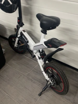 OMW El cykel - Säljer en vit elcykel med svart sadel och detaljer i svart och rött. Cykeln har små hjul med röda kanter, stänkskärmar och ett batteri under sadeln. Styret har flera knappar och reglage samt en hjälm som hänger på. Perfekt för smidig transport i stan.