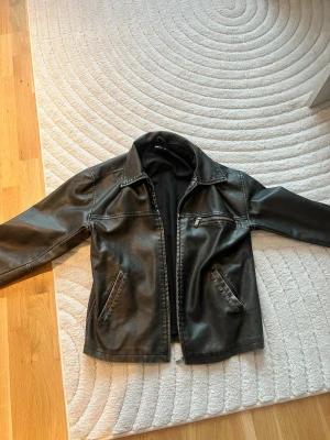 Svart skinnjacka från Gina Tricot - Snygg svart skinnjacka från Gina Tricot i storlek XS/S. Klassisk modell med krage, dragkedja framtill och tre fickor med dragkedja. Perfekt för dig som vill ha en stilren och tidlös jacka till din garderob.