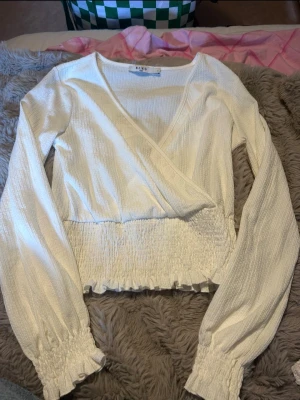 Vit omlottblus med smockdetaljer - Säljer en vit omlottblus med lång ärm och smock i midjan samt vid ärmsluten. Blusen har en djup v-ringning och lätt rynkad struktur i tyget. Perfekt att styla med jeans eller kjol för en trendig look.
