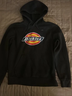 Svart Dickies hoodie med logga - Svart hoodie från Dickies med stor färgglad logga på bröstet. Klassisk modell med huva och känguruficka framtill. Perfekt för en avslappnad streetwear-look.