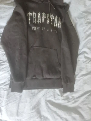 Svart Trapstar hoodie  - Säljer en svart hoodie från Trapstar med stort vitt tryck på bröstet där det står 'Trapstar It's a Secret'. Hoodien har huva med dragsko och en klassisk magficka. Perfekt för dig som gillar streetwear och vill sticka ut.