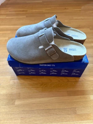 Beige Birkenstock tofflor i mocka - Säljer ett par snygga beige Birkenstock Boston i storlek 43. Som nya och i mycket fint skick utan några brister.  Klassisk och bekväm modell i mjuk mocka med justerbart metallspänne och ergonomisk korkfotbädd. Perfekta både hemma och ute för en avslappnad och stilren look!  Originalkartong följer med. Nypris ca 2000 kr.  Vid bud eller funderingar är det bara att skriva! 😊