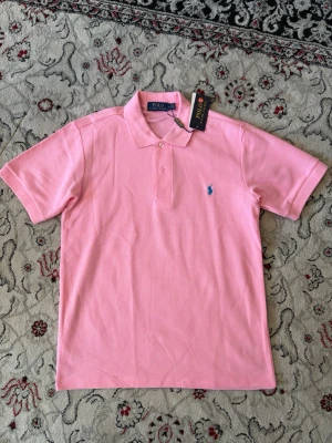 Rosa pikétröja från Polo Ralph Lauren - Snygg rosa pikétröja från Polo Ralph Lauren med klassisk krage och korta ärmar. Tröjan har två knappar framtill och den ikoniska blå loggan broderad på bröstet. Perfekt för en stilren och avslappnad look.