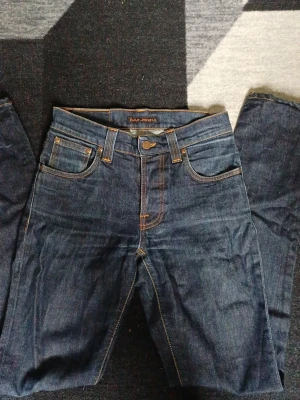 Mörkblå Nudie Jeans raka jeans - Säljer ett par mörkblå jeans från Nudie Jeans med raka ben och klassisk femficksdesign. Jeansen har tydliga orange kontrastsömmar och patchdetalj på bakfickan. Tillverkade i kraftig denim för en stilren look. Skriv vid frågor!