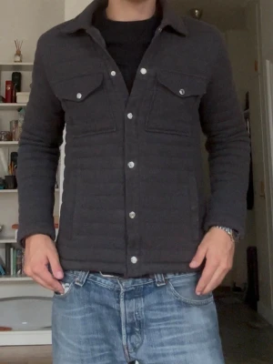 Mörkgrå quiltad overshirt - Säljer en mörkgrå quiltad overshirt med tryckknappar framtill och två bröstfickor med lock. Overshirten har en klassisk krage och raka ärmar. Materialet ser ut att vara mjukt och vadderat, perfekt för lager på lager under kyligare dagar.