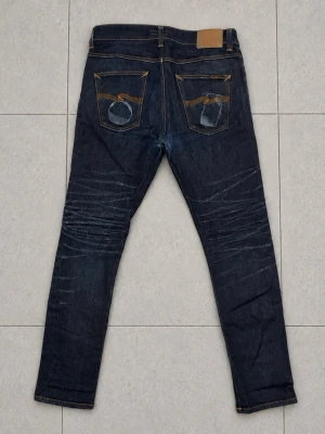 Nudie Jeans 32/32 - Säljer ett par mörkblå jeans med smal passform och snygga slitningar på framsidan. Jeansen har klassiska fem fickor och kontrastsömmar. Bakfickorna har dekorativa detaljer och tvättade effekter. Perfekta för en trendig och avslappnad stil.