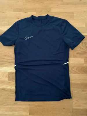 Mörkblå Nike sporttröja - Mörkblå sporttröja från Nike med korta ärmar och rund hals. Tröjan har en vit Nike-logga på bröstet och vita detaljer vid sidorna. Tillverkad i ett lätt och snabbtorkande material som passar perfekt för träning.