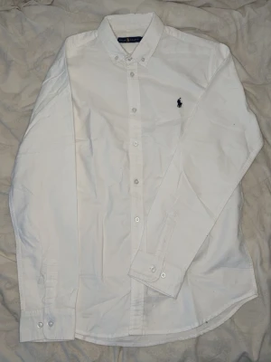 Ralph lauren skjorta - Klassisk vit skjorta från Ralph Lauren i storlek XL. Skjortan har knappar framtill, button-down krage och den ikoniska broderade loggan på bröstet. Tillverkad i bomull och har långa ärmar med manschettknappar. Perfekt för en clean och stilren look.