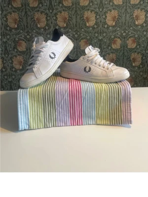 Vita Fred Perry sneakers med svart detalj - Snygga vita sneakers från Fred Perry med svart hälparti och klassisk logga på sidan och bak. Skorna har låga skaft, vita snören och perforerade detaljer på sidorna. Tillverkade i läder med gummisula för en clean och stilren look.