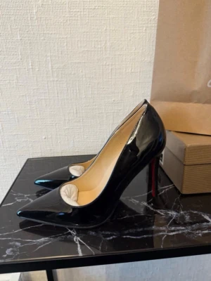 Svarta lackpumps med röd sula - Snygga svarta pumps i lack med spetsig tå och hög klack. Skorna har en ikonisk röd sula och elegant siluett. Perfekta för att lyfta vilken outfit som helst och ger en lyxig känsla. Materialet är glansigt och insidan är ljusbeige.