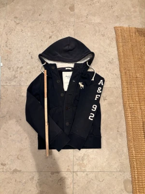 Mörkblå hoodie Abercrombie & Fitch - Mörkblå hoodie från Abercrombie & Fitch med knappar framtill, vit broderad logga på bröstet och texten 'A&F 92' på ärmen. Utrustad med huva och vita dragsnören. Mjuk insida och ribbade muddar. Perfekt för en avslappnad och cool stil. Längd:58cm Axelbredd:40cm