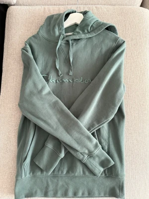 Grön hoodie från Champion - Snygg grön hoodie från Champion med broderad logga på bröstet och liten logga på ärmen. Klassisk känguruficka framtill och justerbar huva med dragsko. Mjuk bomullsmix som är skön att ha på sig. Perfekt för en avslappnad och trendig look.