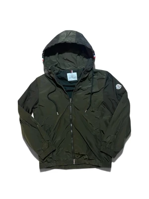 Moncler windbreaker NFC - Stilren mörkgrön vindjacka från Moncler med huva och dragsko, hel dragkedja framtill och elastiska muddar. Jackan har Moncler-logga på ärmen och en diskret röd, vit och blå rand på huvan. Tillverkad i lätt, vattenavvisande material. Jackan har NFC och märket scannas och tar dig till moncler.com