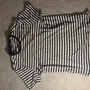 Randig t-shirt med marinblå detaljer - Snygg t-shirt med marinblå och vita ränder samt en liten röd broderad detalj på bröstet. Klassisk passform och rund halsringning med marinblå kant. Perfekt för en avslappnad och stilren look.