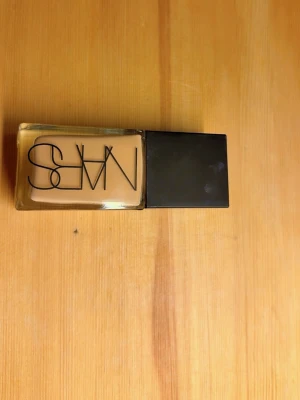 NARS flytande foundation i glasflaska - Flytande foundation från NARS i en rektangulär glasflaska med svart lock. Färgen är ljusbeige och flaskan har tydlig logga på framsidan. Perfekt för en naturlig bas och enkel att applicera tack vare den smidiga förpackningen.