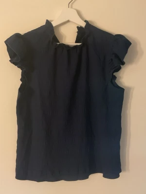 Mörkblå blus med volangärm - Snygg mörkblå blus med volangdetaljer vid ärmarna och lätt struktur i tyget. Blusen har en högre krage och är tillverkad i ett tunt, luftigt material som ger en avslappnad men ändå chic look.