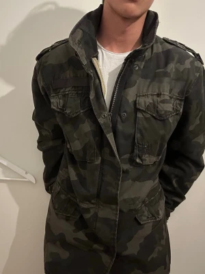 Camo field jacket - Säljer en field jacket i militärgrönt camomönster med flera praktiska fickor och dragkedja framtill. Kan dra till midjan med inbyggda bandet så man får snygg v taper