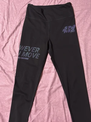 Svarta sportiga leggings med tryck - Säljer ett par svarta leggings med hög midja och sportigt tryck i grått på ena benet och höften. Materialet är stretchigt och passar perfekt för träning eller en aktiv dag. Texten på benet lyder 'NEVER IN MOVE' och på höften står det 'SPORT ACTIVE'.