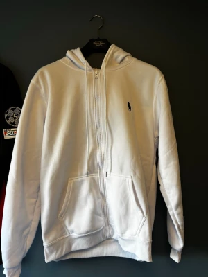 Vit hoodie med dragkedja och huva - Snygg vit hoodie med dragkedja framtill och klassisk huva. Har känguruficka och en liten broderad logga på bröstet. Perfekt för en chill och stilren look. Mjuk och skön bomullskänsla, passar till det mesta i garderoben. Oanvänd säljes då den är för liten för mig. Storlek M.