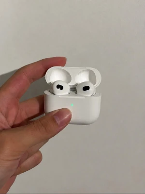 AirPods 3 - Säljer ett par stilrena trådlösa in-ear hörlurar i vitt med laddningsetui. Hörlurarna har silikonproppar och en kompakt, rundad design. Perfekta för musik, poddar och samtal på språng. Laddningsetuiet har en LED-indikator på framsidan.