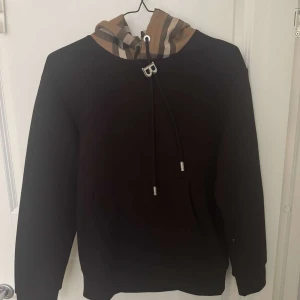 Svart hoodie med rutig huva Burberry - Svart hoodie från Burberry med klassisk rutig huva i beige, svart och vitt. Tröjan har långa ärmar, känguruficka och snörning med metalländar samt ett B-hänge. Perfekt för dig som gillar streetstyle och vill sticka ut med en ikonisk detalj.