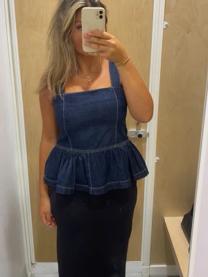 Mörkblå denim peplum-linnetopp - Snygg mörkblå denim peplum-linnetopp med breda axelband och markerade sömmar. Toppen har en figurnära passform upptill och volang nertill som ger en trendig siluett. Perfekt att styla med både kjol och kostymbyxor. Köpt i Spanien. 