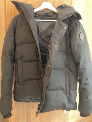 Svart pufferjacka från Canada Goose - Snygg svart pufferjacka från Canada Goose med klassisk patch på ärmen. Jackan har dragkedja och knappar framtill, två stora fickor och en rejäl huva. Perfekt för kalla vinterdagar och riktigt varm tack vare det tjocka materialet.