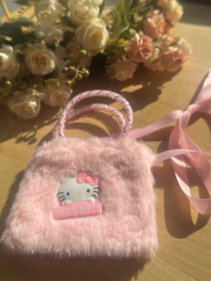 Rosa fluffig Hello Kitty handväska - Supersöt liten handväska i fluffigt rosa material med Hello Kitty-motiv framtill. Väskan har två korta handtag i rosa och ett långt band i matchande färg. Perfekt för dig som älskar gulliga accessoarer och vill sticka ut med en unik look.