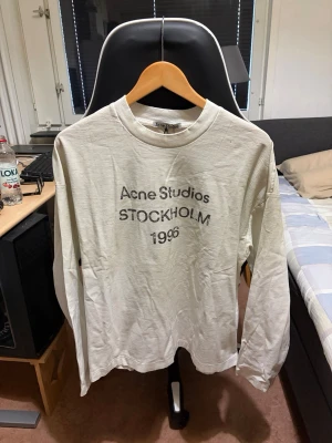 Vit långärmad topp Acne Studios - Vit långärmad topp från Acne Studios med tryck 'Acne Studios STOCKHOLM 1996' på bröstet. Klassisk rund halsringning och avslappnad passform. Perfekt för dig som gillar stilrena och trendiga plagg.