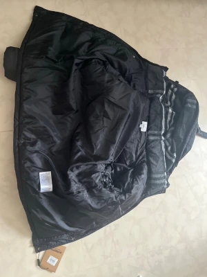 Svart dunjacka från Burberry - Snygg svart dunjacka från Burberry med klassiskt rutmönstrat innerfoder i grått och vitt. Jackan har en vadderad insida som håller dig varm under kalla vinterdagar. Perfekt för dig som vill ha både stil och funktion.