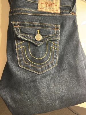 True Religion Joey Low Rise Flare Jeans - Mörkblå jeans från True Religion, modell Joey Low Rise Flare. Snygga kontrastsömmar i gult. Tillverkade i slitstarkt denim med låg midja och utsvängda ben. Jag har tidigare sytt de lågmidjade men sedan sprätt upp vilket syns lite i skrevet men inget man tänker på. Skriv mått eller frågor!💕