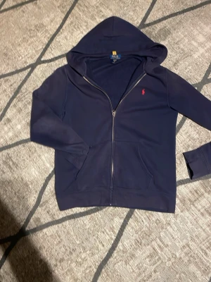 Mörkblå hoodie från Polo Ralph Lauren - Mörkblå hoodie med dragkedja från Polo Ralph Lauren. Klassisk design med huva, två fickor framtill och röd broderad logga på bröstet. Tillverkad i mjukt bomullsmaterial som känns skönt mot huden. Perfekt för en avslappnad och stilren look. Xl barn så skulle säga s vuxen Priset ej satt i sten. Kontakta för frågor!☺️