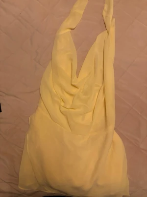 Ljusgul omlottklänning med halterneck - Säljer en ljusgul klänning med halterneck och omlottdetalj. Klänningen har en draperad framsida och är ärmlös, perfekt för sommarens varma dagar. Materialet känns lätt och luftigt, troligen polyester eller liknande.