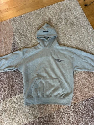 Grå Essentials Fear of God hoodie - Säljer en grå hoodie från Essentials Fear of God med svart logga på bröstet och stor text på ryggen. Hoodien har en stor huva och magficka framtill. Tillverkad i mjukt bomullsmaterial, perfekt för en avslappnad och trendig look.