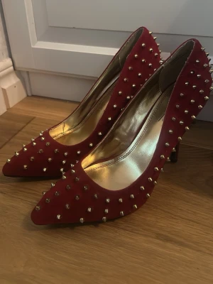 Röda pumps med nitar från Anne Michelle - Snygga röda pumps från Anne Michelle med spetsig tå och täckt klack. Skorna är dekorerade med guldfärgade nitar över hela ovandelen och har en insida i metallic guld. Materialet ser ut att vara mockaimitation. Säljer pga dom är förstora för mig